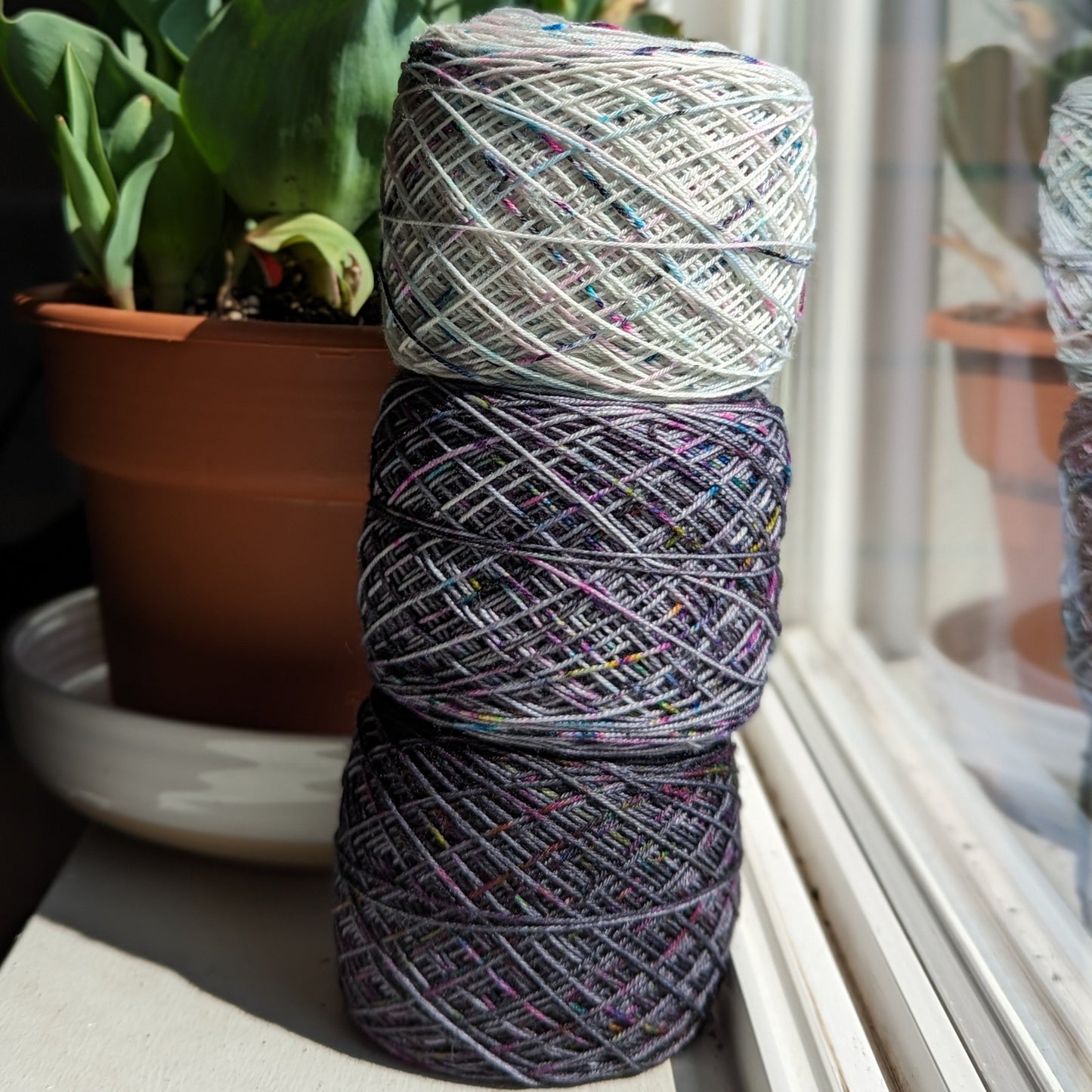 Three stacked skeins of variegated yarn in gray, purple, and black with multicolored flecks, placed on a windowsill next to a potted tulip.